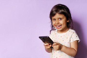 Fototapeta premium Photo of smiling child girl with dark hair using mobile phone and looking at camera isolated on purple background