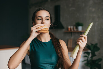 Fototapeta premium Hungry woman eating burger, holding vegetable, standing in the kitchen. Choosing between healthy and unhealthy food. Sporty young girl. Holding hands. Fast food