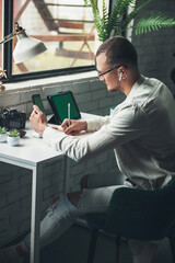 Man entrepreneur in eyeglasses sitting and working at table, writing in planner holding smartphone and using tablet. Creates, planning to-do list in personal