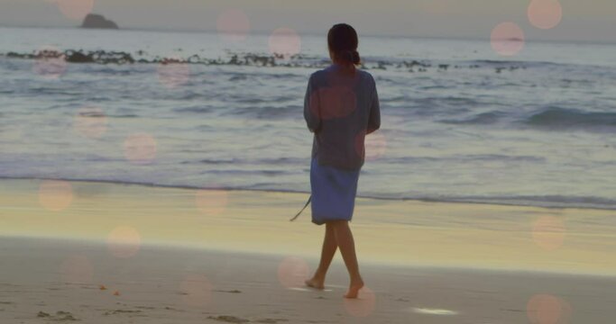 Animation of light spots over biracial woman walking on beach