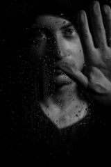 Black and white portrait of a young hooded man holding his hand on a glass covered in water drops - focus on the glass with the drops and his hand