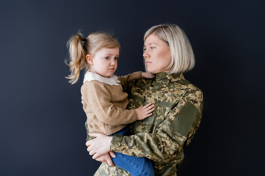 woman in camouflage holding upset and frowning daughter isolated on black.