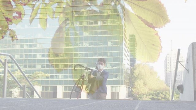 Animation of leaves over asian man with face mask carrying bicycle