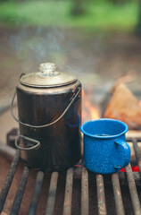 A pot of water and a mug warm up on a metal grate over a fire at a campground in the summer.