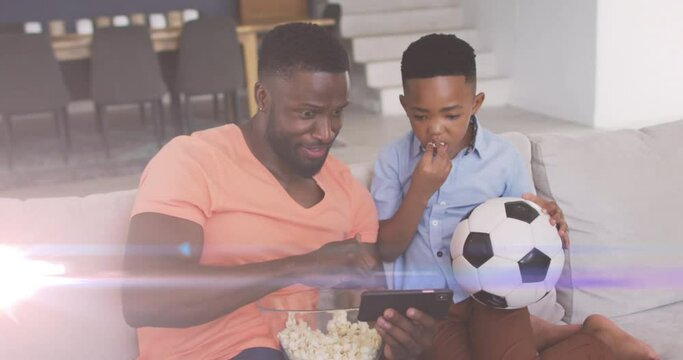 Animation of lights over happy african american father and son watching soccer match on smartphone
