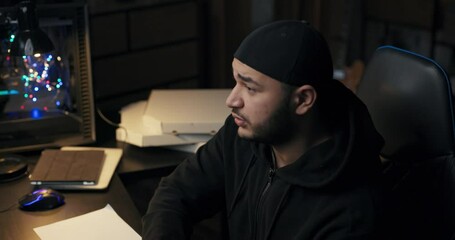 Tired, sleepy, overworked man in cap and sweatshirt works at night in dark room, sleeps in front of computer with head on desk over notes, wakes up suddenly rubs eyes looks into computer screen. - Powered by Adobe