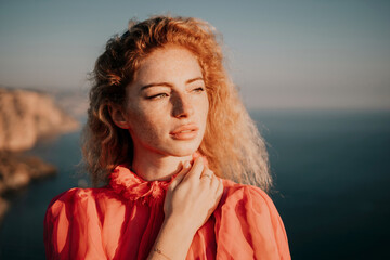 Close up shot of beautiful young caucasian woman with curly blond hair and freckles looking at camera and smiling. Cute woman portrait in a pink long dress posing on a volcanic rock high above the sea