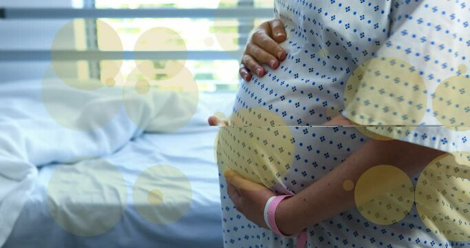 Animation of green spots over biracial pregnant woman touching her stomach in hospital
