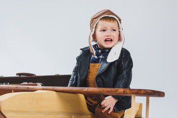 The boy is crying in a leather jacket and a pilot's hat, a wooden plane, brown suitcases. Children's wooden toys. Baby emotions