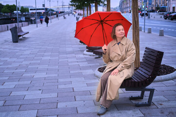 A woman with a red umbrella sits on a city street bench