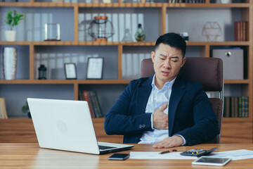 Young Asian man in the workplace is experiencing chest pain, heart attack, stroke. Holds on to his chest, difficulty breathing, wincing in pain. In the office, sitting at a table