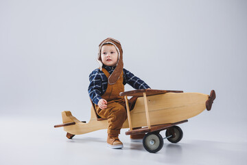Little boy 3 years old plays with a wooden plane on a white background. In overalls and a pilot's hat. Children's eco toys made of natural wood. wooden plane