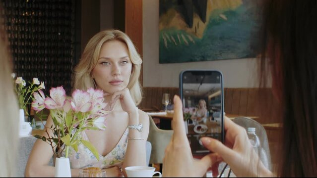 A young blonde woman is sitting in a cafe and drinking coffee. Unrecognizable female hands take pictures of a posing girl in a cafe on a phone camera. A woman poses on a phone camera
