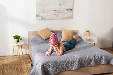Obraz premium young woman with pink hair in bra and jeans using smartphone while lying on bed.