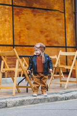 Fototapeta premium Stylish boy 3 years old in a leather jacket and brown trousers sits on the terrace near the cafe. Modern child. Children's fashion. Happy child
