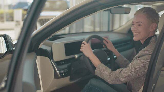 Close-up of happy female testing buying car from inside during visit auto dealership. Test driving of new generation electric vehicle with self driving system. Woman sitting behind the wheel of car.