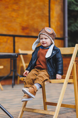 Stylish boy 3 years old in a leather jacket and brown trousers sits on the terrace near the cafe. Modern child. Children's fashion. Happy child