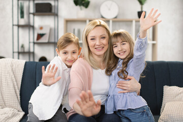 Happy mom and two little sibling kids enjoying leisure at home together, relaxing on couch, hugging, waving their hands. Loving mother embracing sweet children with care, affection. Family concept