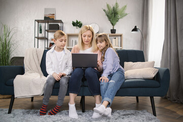 Single Caucasian mother and little children sitting on couch using laptop, family enjoy distant talk by videocall, spend time in internet buying, e-commerce users, cartoons or video fun concept