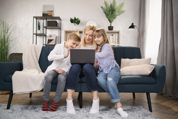 Happy family with two children having good time using laptop. Young smiling mother with cute daughter and son sitting on sofa at home looking at computer screen and watching video.
