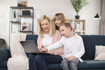 Happy family of mother, little son and daughter sitting together on comfortable couch, using laptop, holding video call with father or greeting grandparents, recording video in living room.