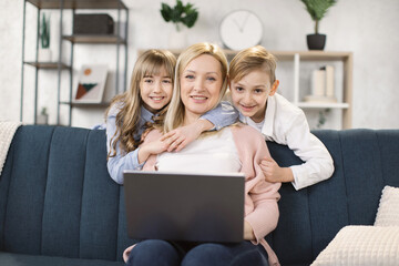 Happy young caucasian mother relax on comfortable couch with little kids have fun watch video on laptop together, charming family rest on sofa in living room using computer with children