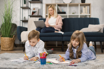 Fototapeta premium Family, leisure and people concept - happy mother with tablet pc computer laptop sitting on sofa and looking at daughter and son drawing at home