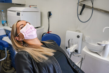 Young woman sitting in dentist chair, mouth still covered with pink face mask, looking into camera