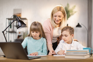 Fototapeta premium Dughter and son doing school lessons together looking at a laptop monitor. Mother stands behind and watches and shows the task on paper.