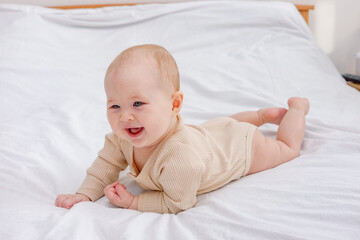 A cute blue-eyed and fair-haired caucasian baby laying on his tummy on white linen on the bed and laughing. Baby care, nursery, developing, parenthood concept.