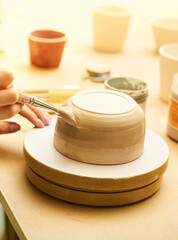 Close-up of girl painting clay mug with glaze. Woman coloring pottery in workshop with a paintbrush. Painter in green apron glazing clay pot.
