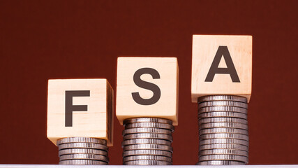 text FSA written on wooden blocks with stacked coins on brown background