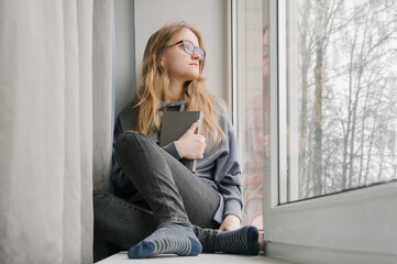 A beautiful young woman with glasses is sitting on the windowsill with a book. Rest at home.