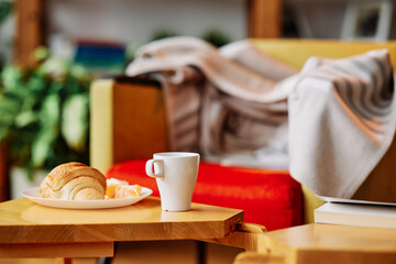 A breakfast pastry on the plate and a cup of coffee are on the wooden table in the living room.