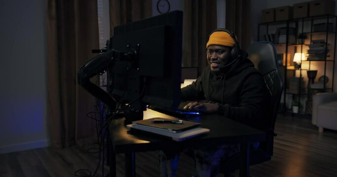 A man's success in a computer game. Boy in sweatshirt cap with headset raises hands up showing strength, victory, winning. He is smiling happy, hanging out in a dark room with led lights.