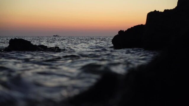 Sea waves splash among the rocks, in the background a boat sailing against the background of the predawn sky