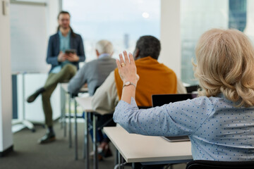A senior woman is asking for an explanation in the computer course while sitting with elderly students in the classroom.