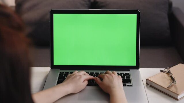 Rear view of a woman typing on the laptop with green screen, sitting at the wooden table. Female working at his desk at the computer, chromakey. Woman uses laptop with green mock-up screen.