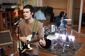 Portrait of young man singing to microphone and playing guitar while composing music with band in...