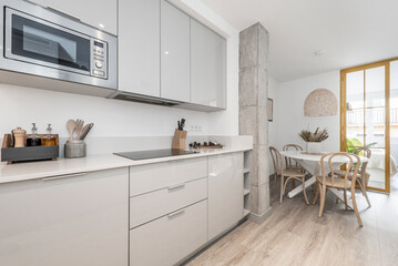 Kitchen with gray wooden furniture combined with white stone countertop and small appliances, cabinets, ceramic hob and microwave oven and circular white wooden dining table with wooden chairs