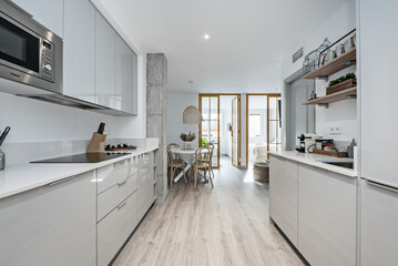 Open kitchen with bright wooden furniture combined with white stone countertop and appliances, dining room with round wooden table and windows in the background