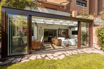 Sunroom with wide open sliding doors with wooden and wicker furniture inside and a stone tile walkway and grassed garden