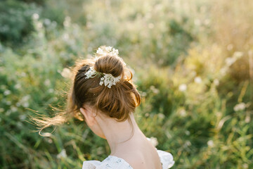 Tender little girl wearing natural white dress with wildflower motiv with wild carrot flowers in hair standing in the field at summer, outdoor lifestyle backlit photo. People from behind
