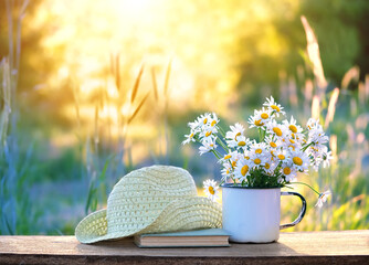 chamomile flowers in cup, book, braided rustic hat on table in garden, sunny natural abstract...