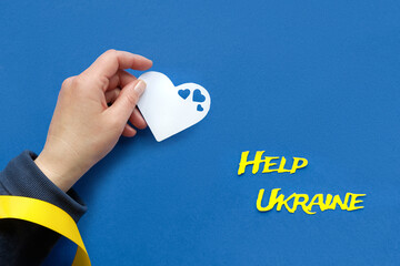 Text Help Ukraine, hand with yellow ribbon holds heart paper silhouette. Volunteer help to refugees...