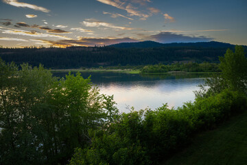 Fototapeta na wymiar 2022-04-11 COEUR D'ALENE RIVER NEAR HARRISON IDAHO WITH A NICE REFLECTION AND BRIGHT GREEN TREES IN THE FOREGROUND WITH A MOUNTAIN RANGE AND NICE SKY AT DUSK