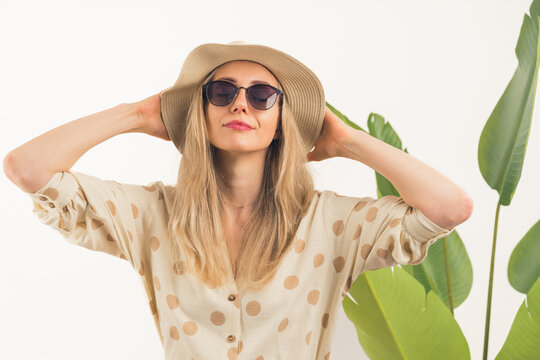fascinating Caucasian blonde girl with sunglasses and hands on her beige hat standing in front of camera medium closeup white background with green leaves studio shot copy space. High quality photo