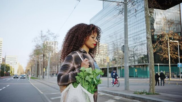 Long-haired curly woman wears glasses and denim overalls, walking in the summer city. Mixed race girl crosses the street on the way from the market with a bag of greens on her shoulder