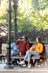 Obraz na płótnie Canvas Happy tourist looking through binoculars near girlfriend with coffee to go sitting on bench outdoors.