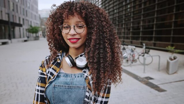 A beautiful African American ethnicity girl with headphones around her neck stands in the middle of a modern city, a young student